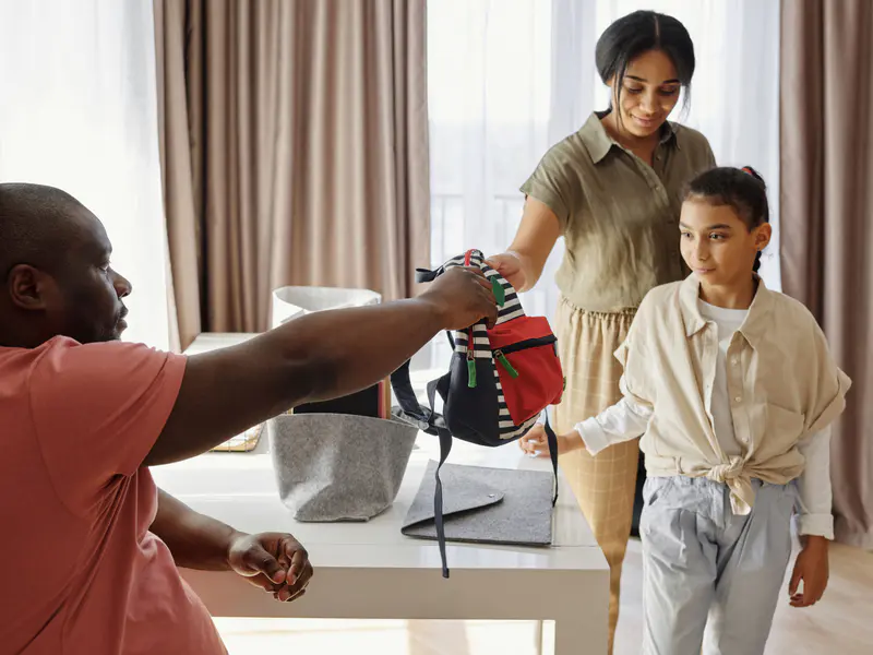 Parents handing a school backpack to their daughter at home — family, schools and health insurance