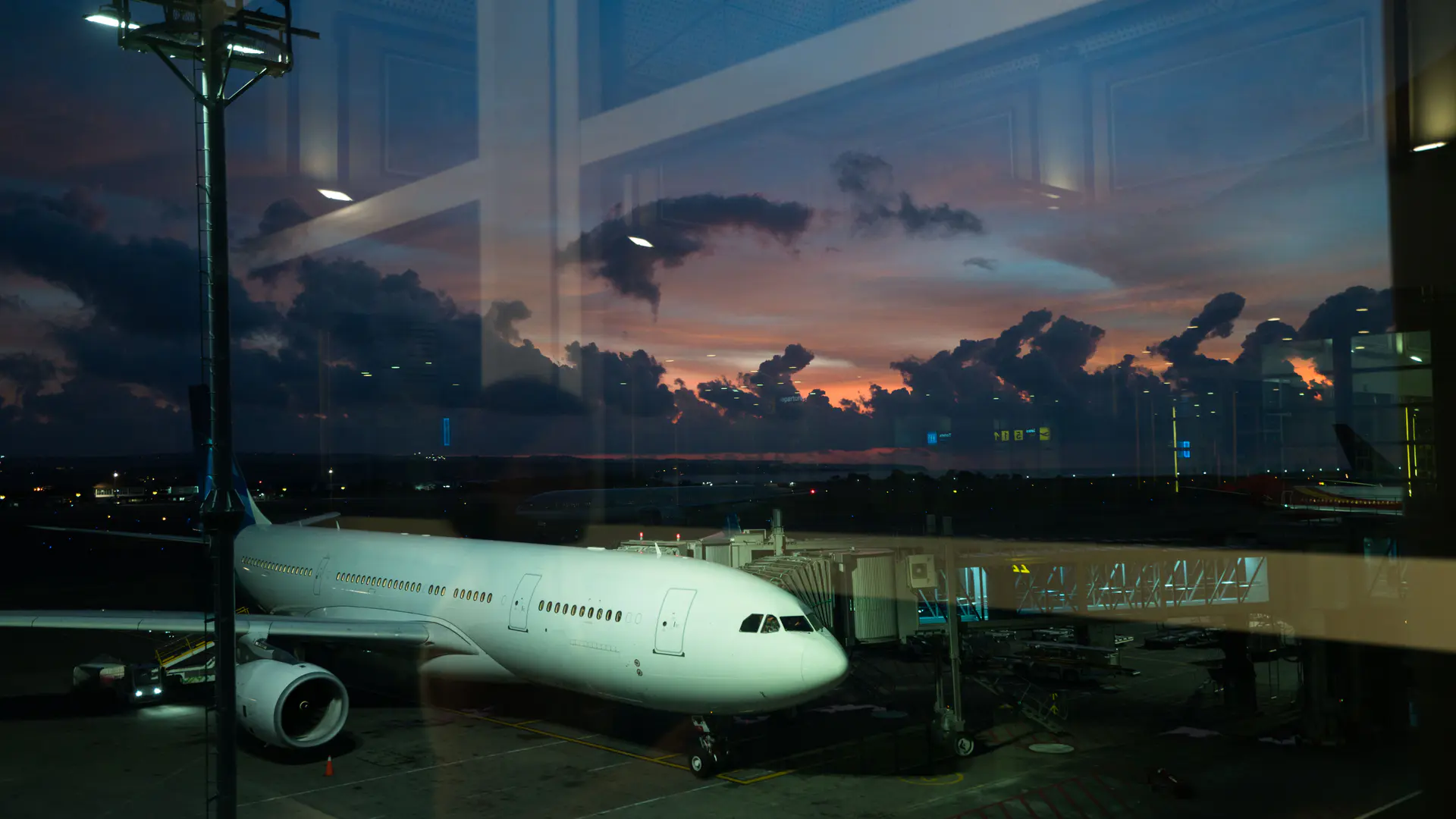 Wide-body aircraft waiting at a boarding bridge through terminal glass at dusk